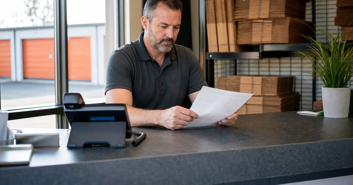 Storage facility office counter with a landline phone, a manager looking at a paper list, sunlit window and a rack of moving boxes behind